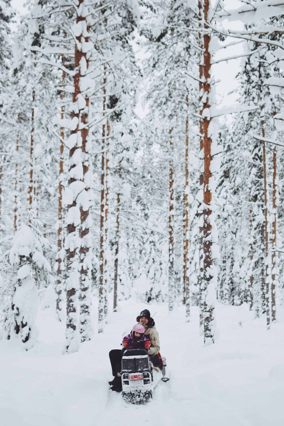 Fredrik is sitting on a snowmobile together with their daughter Lo. The snow lays thick both on the ground and the tree branches.