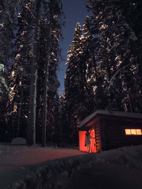A person stands in the doorway of a timbered cottage. The snow is thick on the ground and pines rise high against the dark sky.