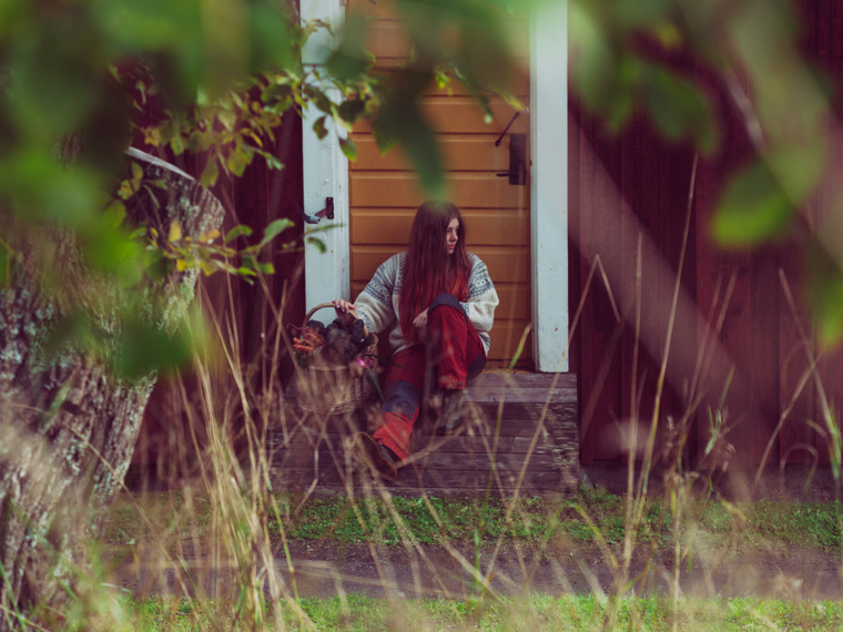 Thea is sitting outside, with her back leaning against the door of a small cottage.