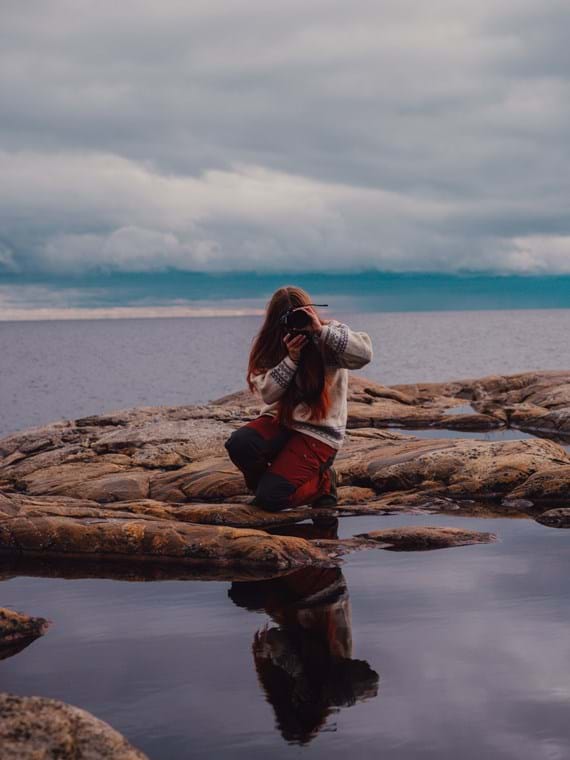På stenhällarna med havet i bakgrunden står Thea, hukad med ett knä i marken och kameran riktad rakt mot fotografen.
