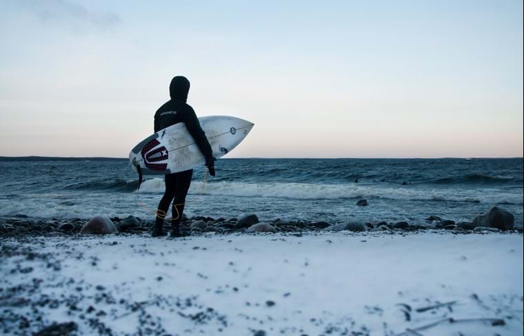 On a lightly snow-covered beach, Klas is standing with his surfboard under his arm and gazing at the horizon