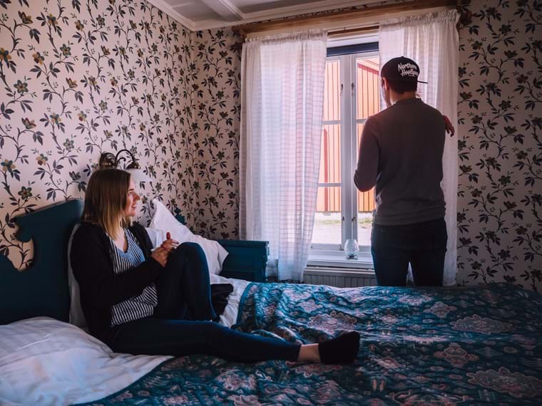 Isabelle is sitting on the bed that is made of floral sheets, her partner Fredrik is standing with their daughter Lo in his arms looking out the window.