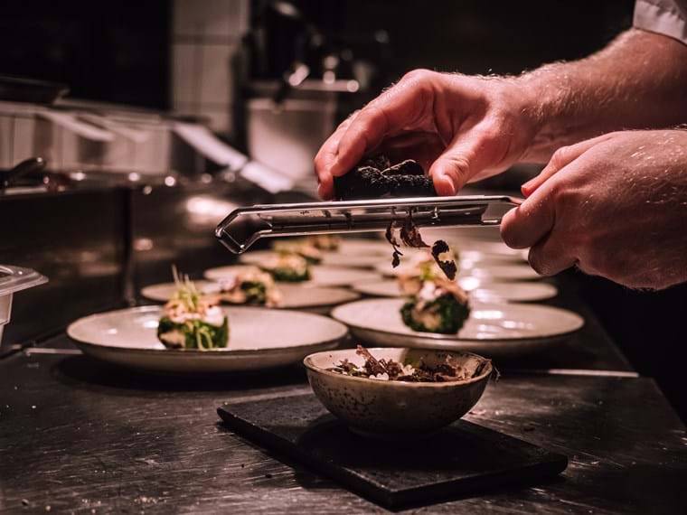 Hands using a grater to grate truffles over a finished dish, in the background are rows of plates with ready-made dishes.