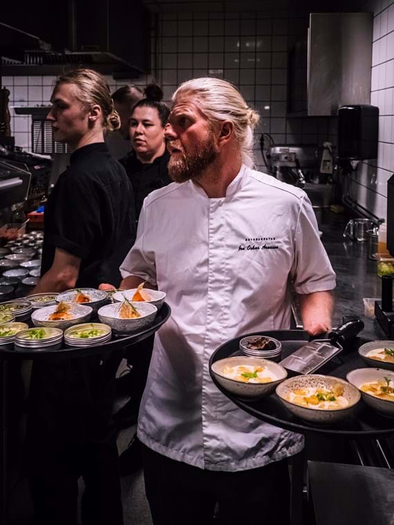 Jon Oskar stands in the kitchen with a tray in each hand that is filled with small bowls containing beautiful dishes. In the background more chefs can be seen, ready to do the next dish.