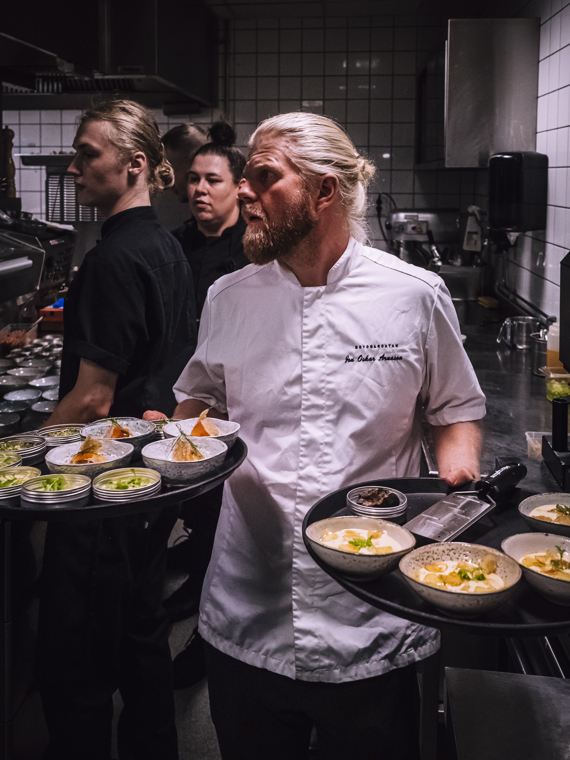 Jon Oskar står i köket med en bricka i varje hand som är fyllda med små skålar innehållande vackert upplagda rätter. I bakgrunden syns fler kockar, redo att lägga nästa rätt.