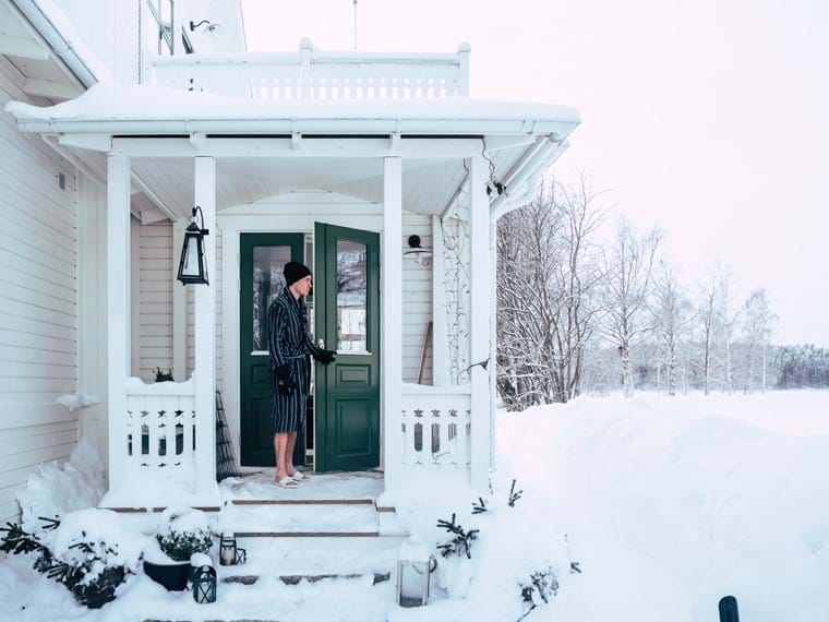 John stands on the porch of the beautiful 20's house wearing a hat, bathrobe, and slippers. The snow is thick on the ground.
