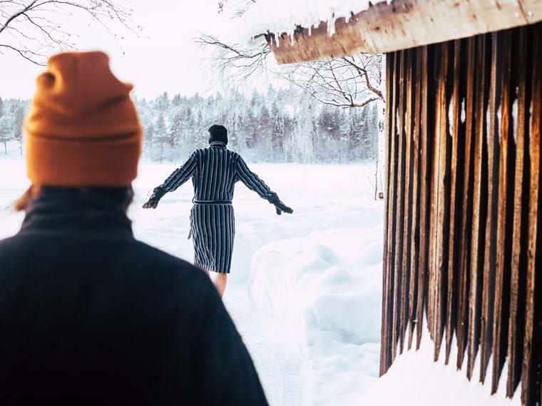 : Maia in the foreground wearing a hat and bathrobe, watches as John follows the passage past the sauna down to the water.