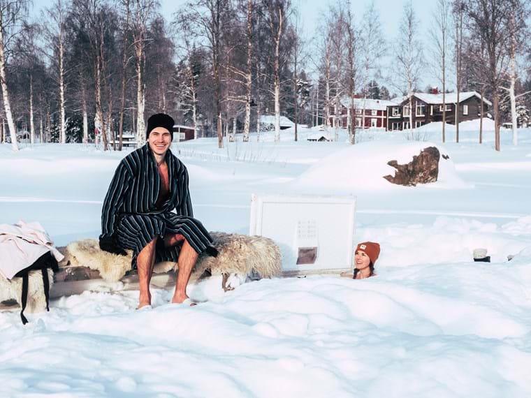 A happy John wearing a bathrobe and hat, is sitting on a bench covered with sheepskin on the ice. Next to him, Maia takes a dip in the ice wake.