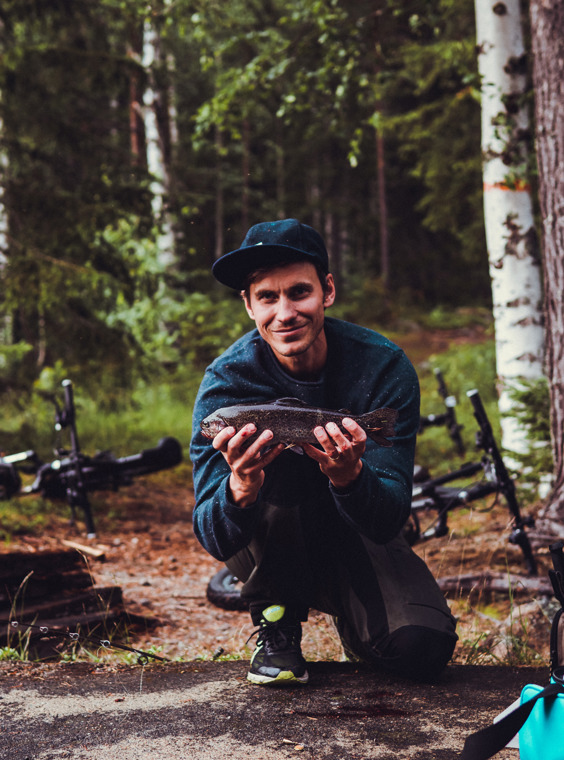 John stands crouched with one knee on the ground and proudly shows off the fish.